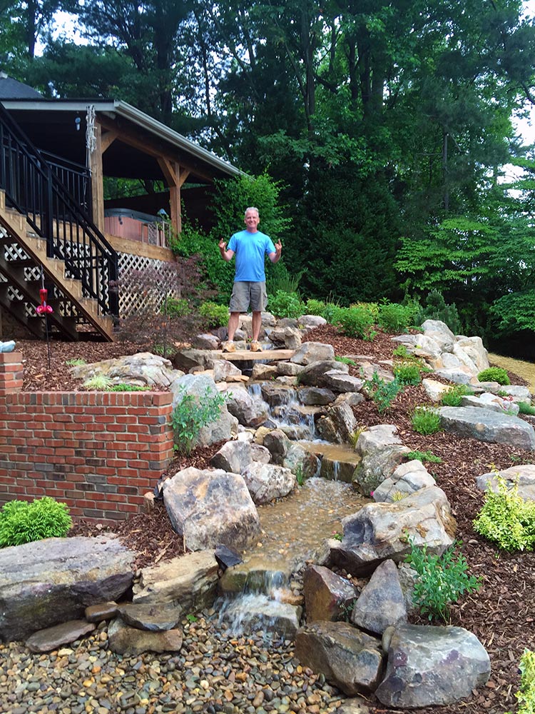 Man standing by backyard waterfall and landscaped garden.